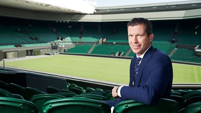 Tim Henman wearing a blue blazer. he's sitting on green chairs looking back and smiling to the camera. Wimbledon centre court in the background