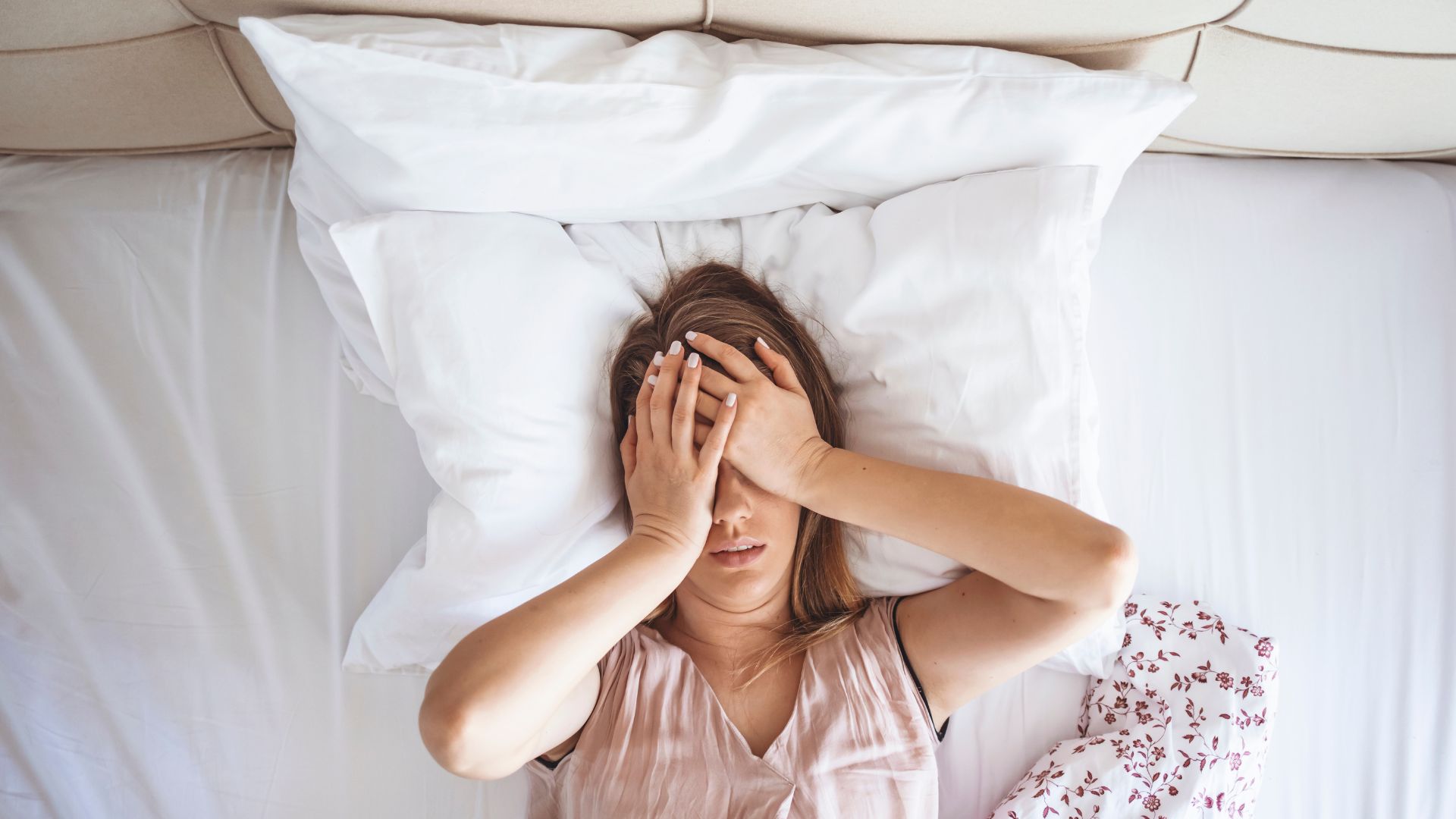The image shows a sleep deprived woman lying in bed and holding her hands over her eyes