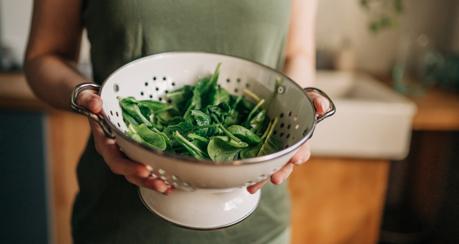A woman in a khaki green sleeveless top holds a white colander full of freshly washed raw spinach, a magnesium rich food that has multiple benefits to a person's health and sleep