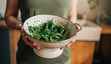 A woman in a khaki green sleeveless top holds a white colander full of freshly washed raw spinach, a magnesium rich food that has multiple benefits to a person's health and sleep