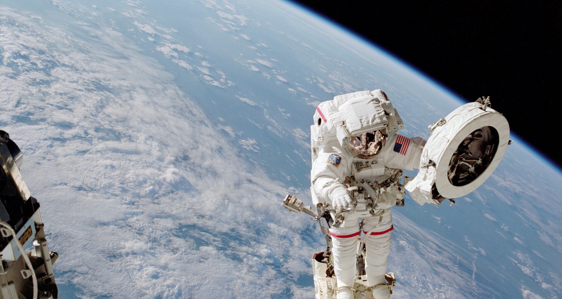An astronaut in a white spacesuit works on a fixture while outside of the International Space Station. He holds a large white circular object in his left hand. Earth can be seen behind him: the planet's blue water and white clouds take up most of the background, but a sliver of black space can be seen at top right.