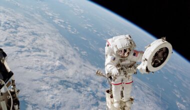 An astronaut in a white spacesuit works on a fixture while outside of the International Space Station. He holds a large white circular object in his left hand. Earth can be seen behind him: the planet's blue water and white clouds take up most of the background, but a sliver of black space can be seen at top right.