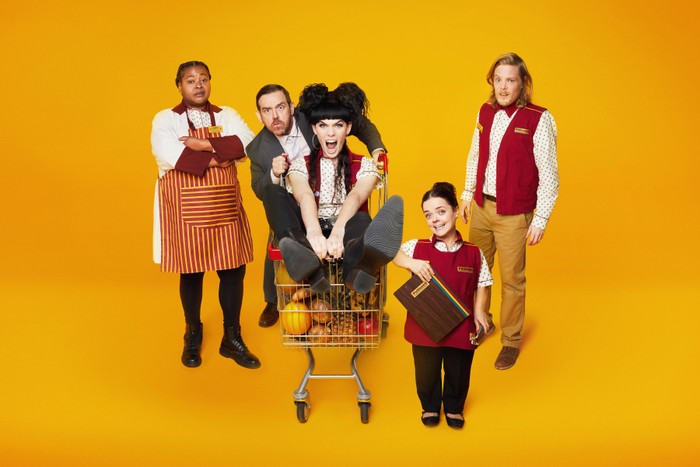 Kayla Meikle as Beefy Linda, Nick Frost as Simon, Jordan Gray as Liv, Francesca Mills as Millie and Thomas Gray as Tom in Transaction. They are stood in front of a yellow background and Millie is sat in a shopping trolly.