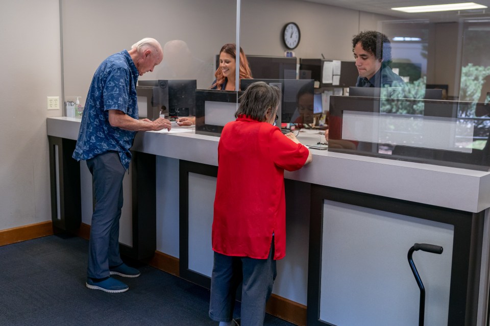 Two Latinx customer service representatives assisting senior customers at a bank or medical office reception desk.