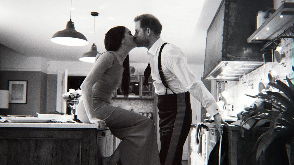 Black and white still image of Prince Harry and Meghan Markle kissing in a kitchen.