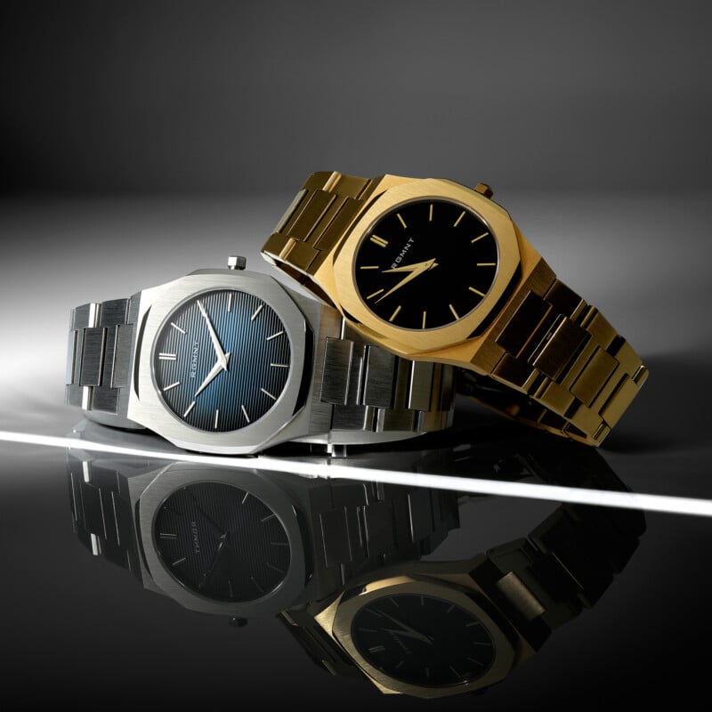Two luxury wristwatches, one silver with a blue dial and one gold with a black dial, are displayed side by side on a reflective black surface with dramatic lighting and shadows.