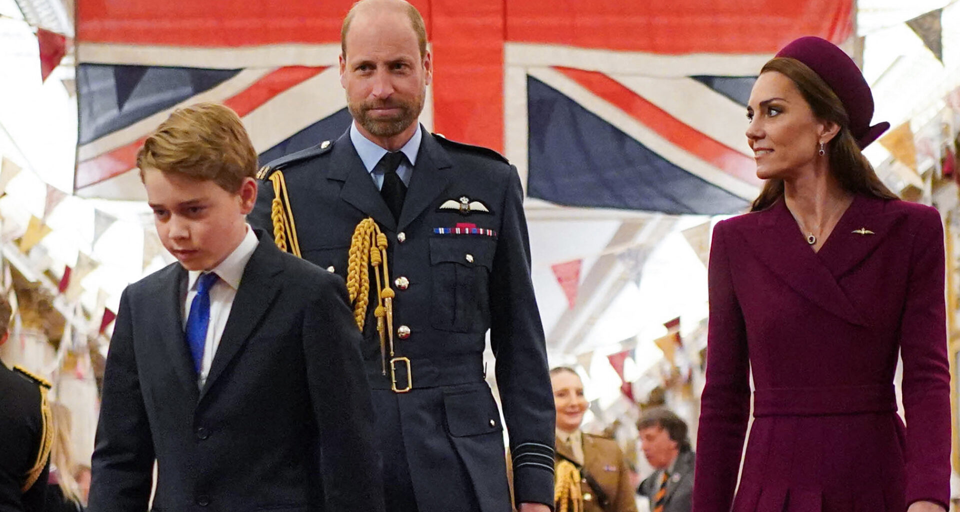 Kate Middleton looks at Prince William, who is wearing military uniform in front of the British flag, while Prince George walks with his head down