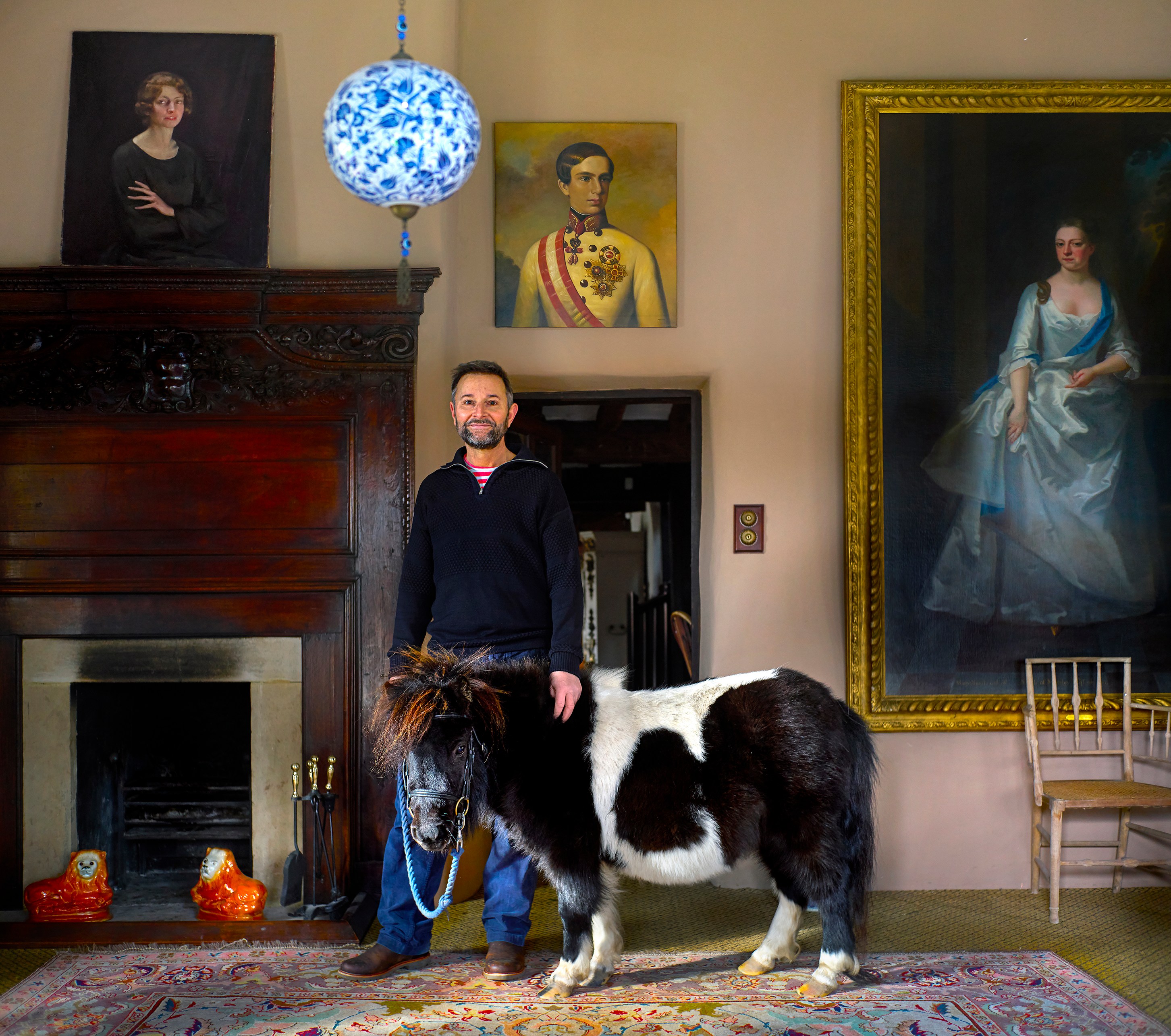 A man stands with his miniature horse in a room with antique paintings and a fireplace.