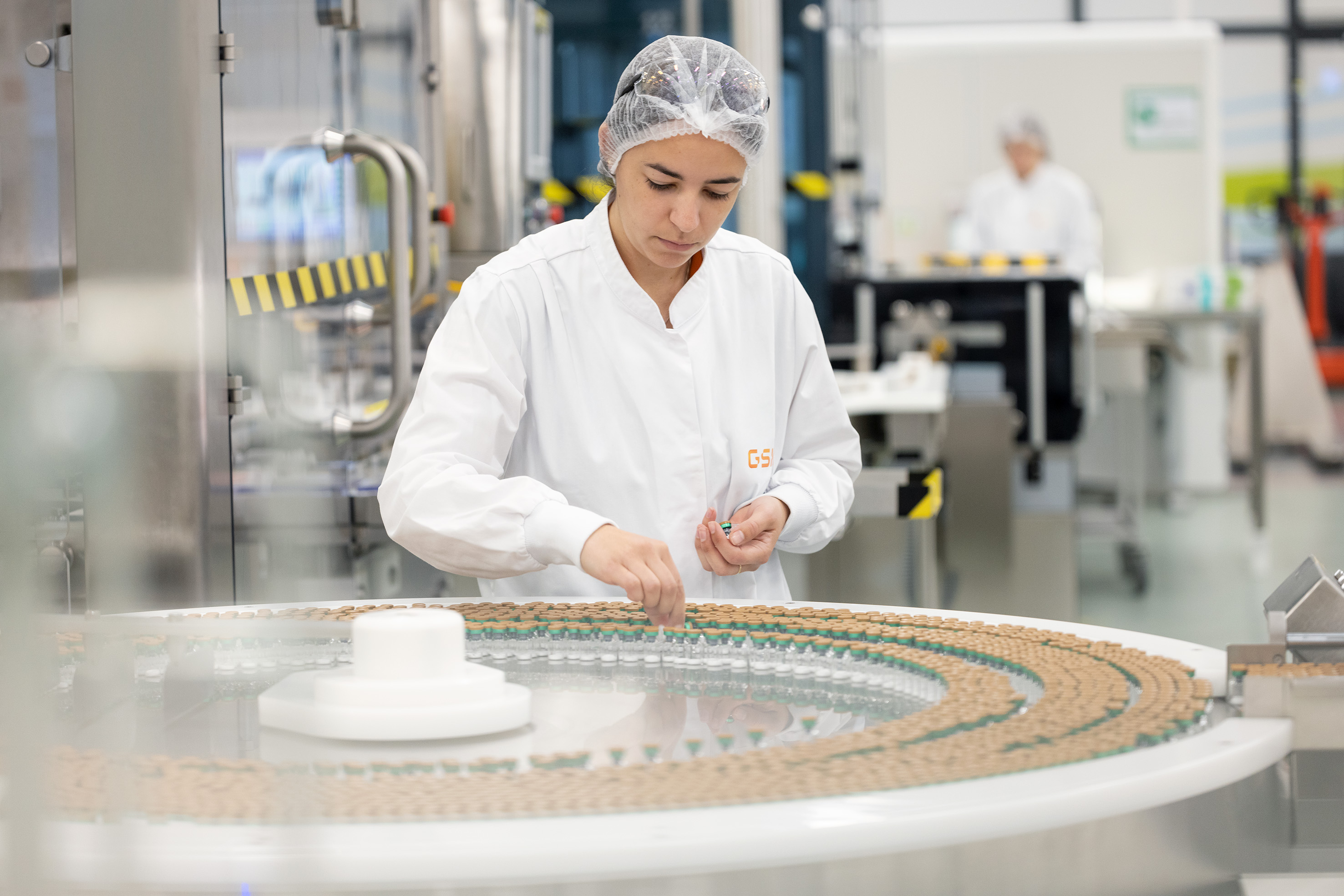 A worker in a lab coat placing vials on a conveyor belt.