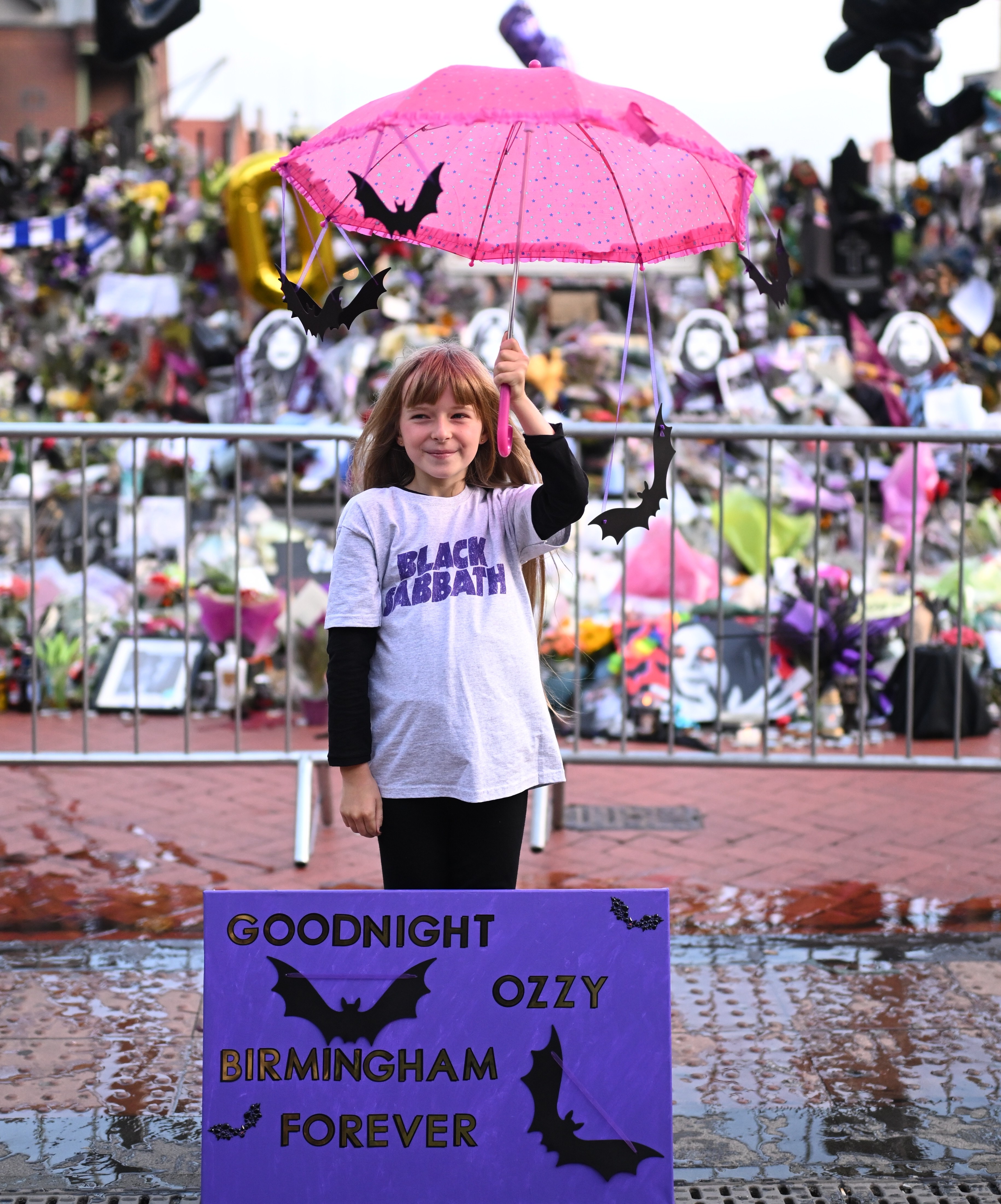 Girl holding a pink umbrella with bat decorations and a sign that reads "Goodnight Ozzy Birmingham Forever" in front of a tribute to Ozzy Osbourne.