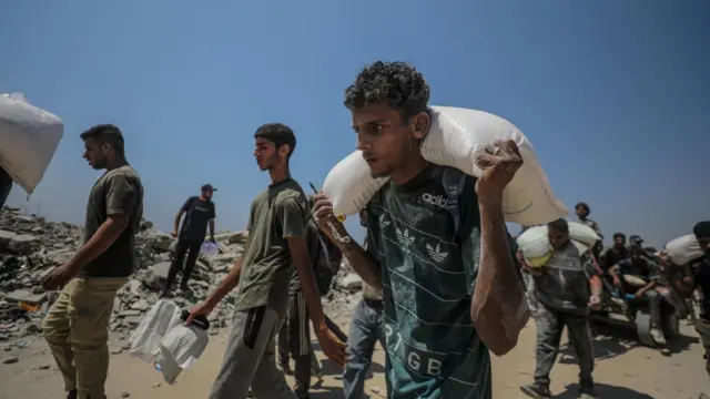 A group f men carry white sacks containing aid down a dusty road past destroyed buildings