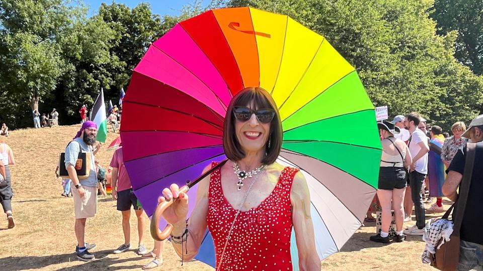 One person holds a huge rainbow umbrella and smiles into the camera. They are wearing large sunglasses and a sparkly red dress. Behind are lots of people in the park, some with flags, and trees behind them