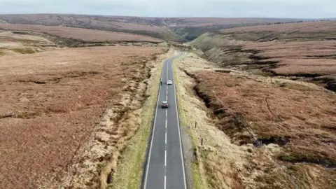 BBC The Snake Pass road winds through open countryside. There are two cars on the road and a cyclist.