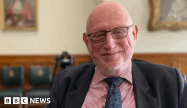 A man in a suit jacket, blue tie and red and white striped shirt smiles at the camera. He is bald, with a grey beard and glasses.