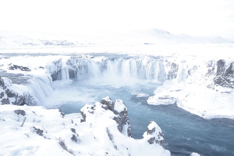 A wide waterfall cascades into a blue river surrounded by snow-covered rocks and a white, wintry landscape under a bright, overcast sky.