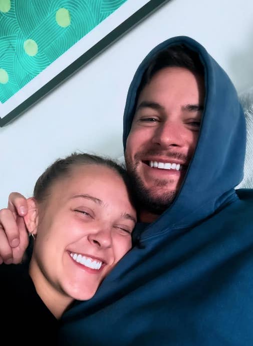 Two people smiling closely, one wearing a hooded sweatshirt. They appear happy and relaxed