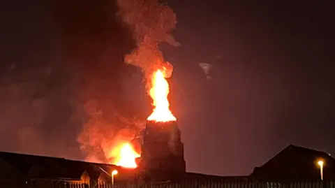 BBC Flames are coming out of a tower, silhouetted against a dark sky. You can see some rooftops and street lights.