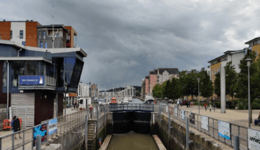 The lock gates at Portishead Marina are in the centre of the image showing the water is it holding. There are many boats and buildings in the background and people walking along the marina.. The sky is overcast.