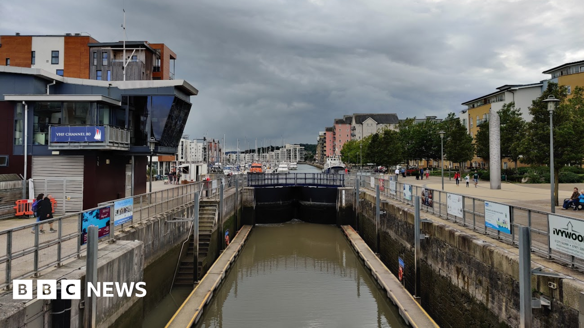 The lock gates at Portishead Marina are in the centre of the image showing the water is it holding. There are many boats and buildings in the background and people walking along the marina.. The sky is overcast.