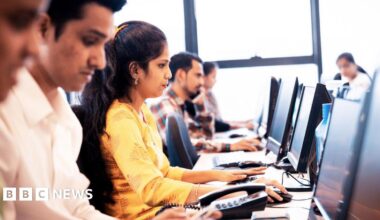The image shows Indian workers at a software company seated in front of their computer screens. A woman wearing a white kurta is prominently seen, surrounded by male colleagues on either side.