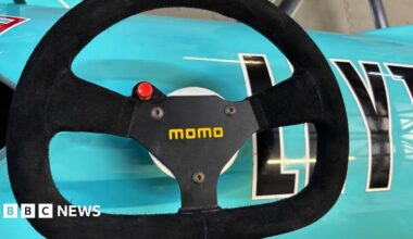 A black steering wheel with a red button and yellow font saying "momo" on it. The wheel is placed on a turquoise car.