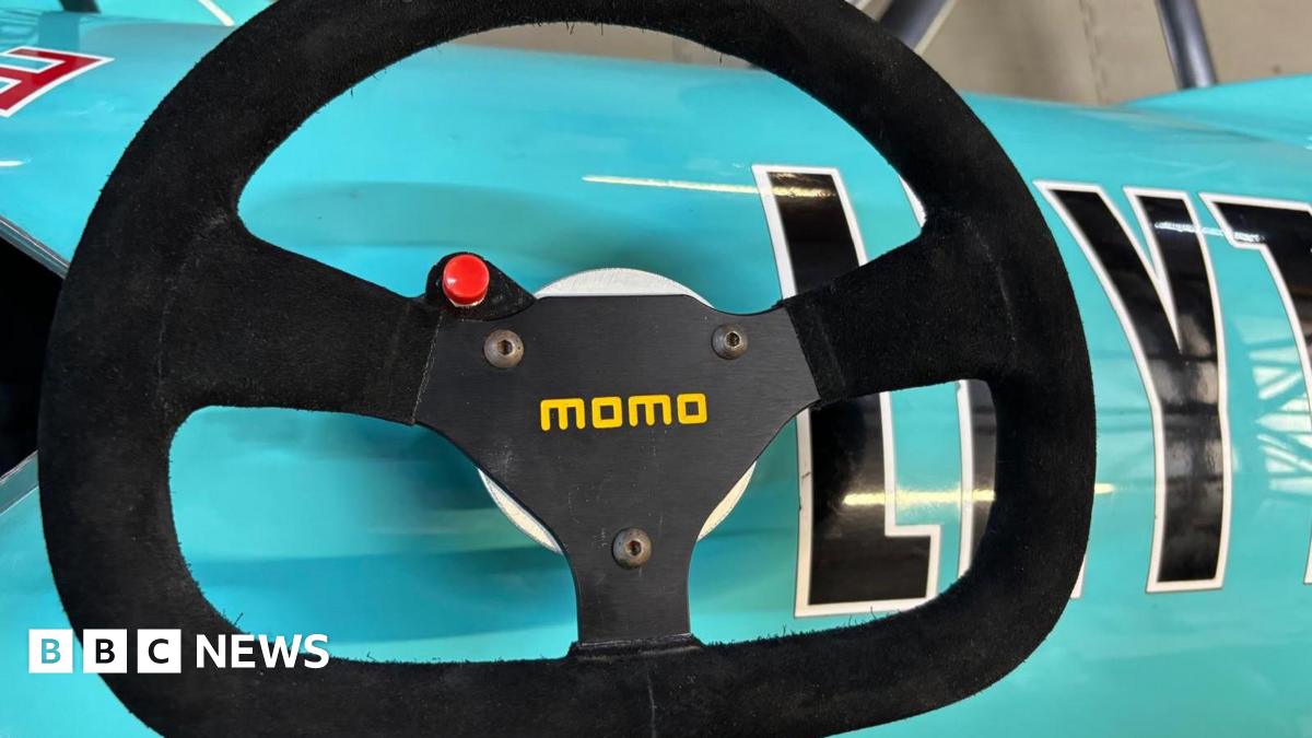 A black steering wheel with a red button and yellow font saying "momo" on it. The wheel is placed on a turquoise car.