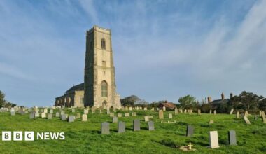 A 15th Century church surrounded by its graveyard. There are many gravestones, and there are some houses next to one of its boundaries.