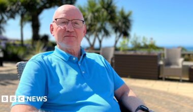 Mike is sitting outdoors on a sunny day, wearing a blue polo shirt and dark pants. The background includes palm trees, lush greenery, and outdoor furniture under a clear blue sky.