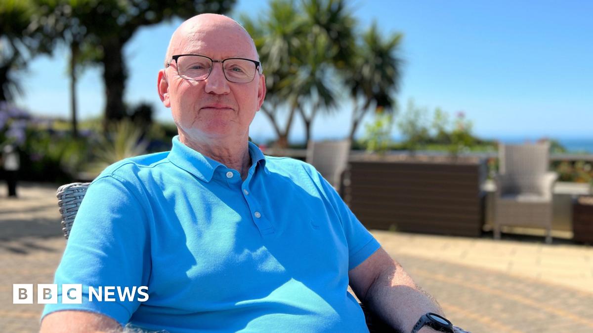 Mike is sitting outdoors on a sunny day, wearing a blue polo shirt and dark pants. The background includes palm trees, lush greenery, and outdoor furniture under a clear blue sky.