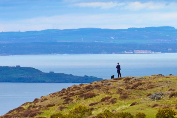 A man and his dog stand atop Arthur's Seat in Edinburgh, Scotland, overlooking the Firth of Forth as dramatic clouds roll in.