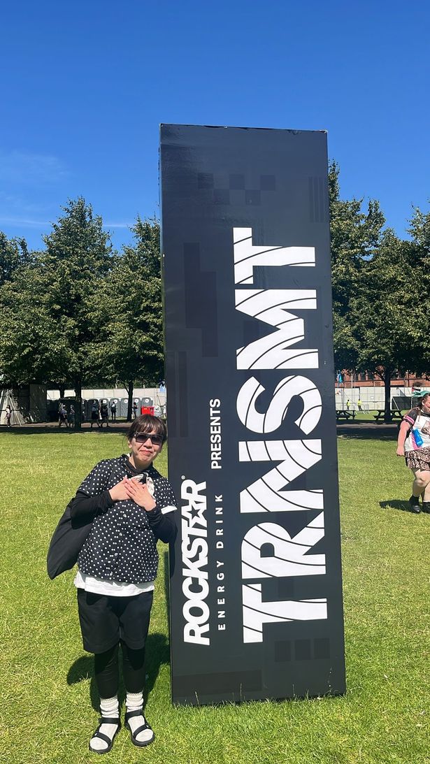 An image of a dark-haired woman with sunglasses, wearing a black top and trousers, standing next to a black pillar reading "Rockstar energy drink presents TRNSMT"