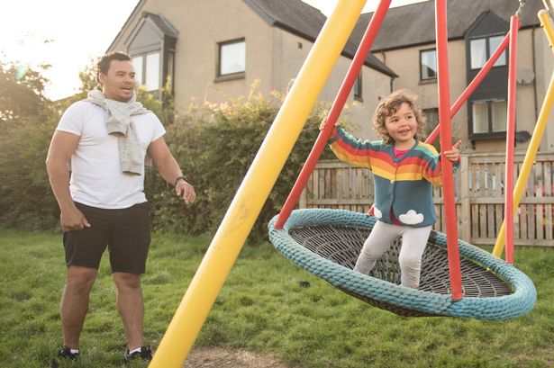 A father plays with her daughter on a swing in a public play park in a sunny day in Edinburgh, Scotland, United Kingdom