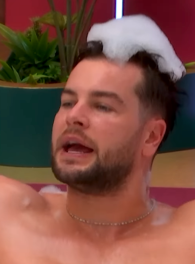 A person with short hair is in a bubble-filled bath, speaking or singing. Background shows decorative plants