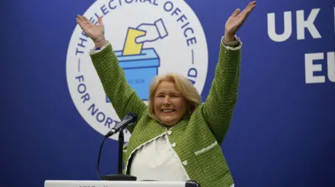 PA Media Pat Cullen throws her hands in the air and smiles as she becomes elected to Westminster in July 2024's UK general election. She has shoulder-length blonde hair, and is wearing a green coat with white stitching and heart-shaped buttons. She is also wearing a white shirt underneath. 