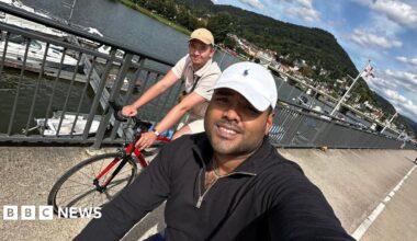Ziyad Azad and Aaron Moloney cycling along the waterfront with boats and a large hill behind them. Mr Azad is taking the selfie photo while cycling and wearing a white hat and black fleece. Mr Moloney is seen in the background wearing a beige cap and an off-white polo top.