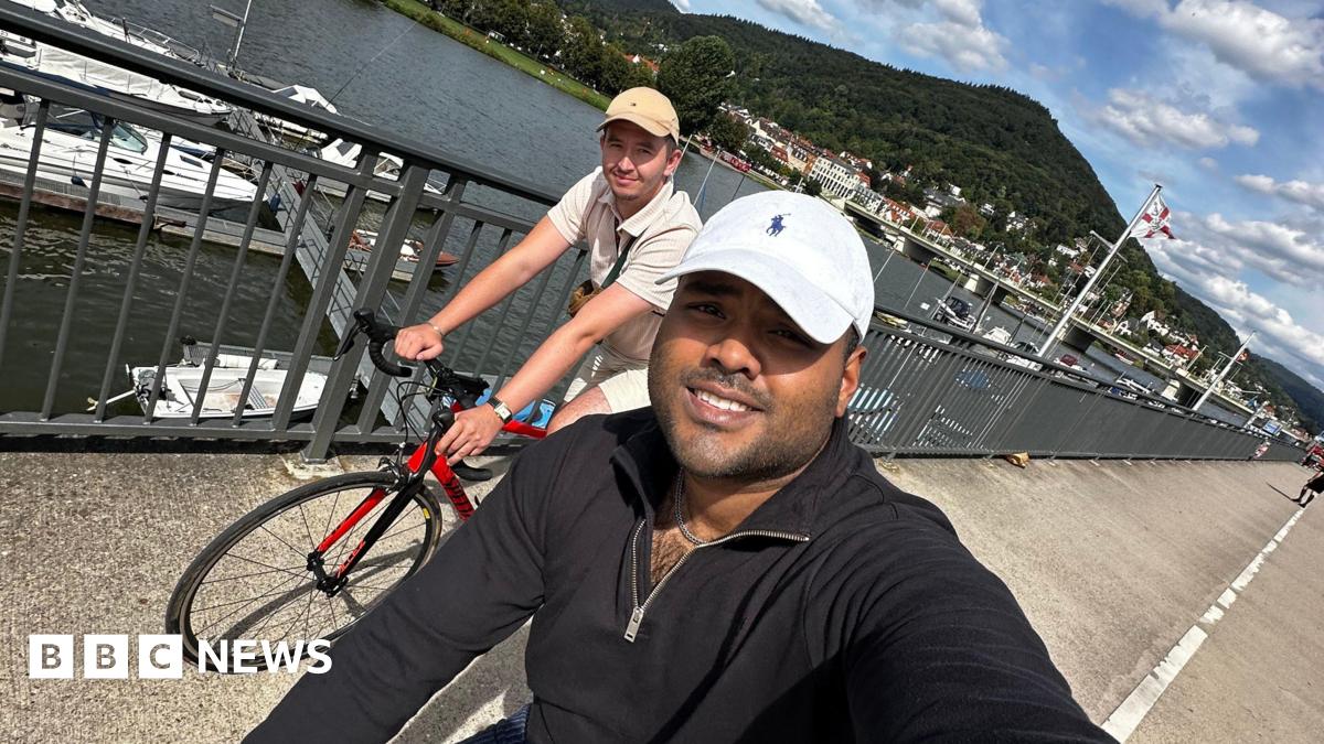 Ziyad Azad and Aaron Moloney cycling along the waterfront with boats and a large hill behind them. Mr Azad is taking the selfie photo while cycling and wearing a white hat and black fleece. Mr Moloney is seen in the background wearing a beige cap and an off-white polo top.