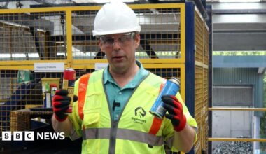 A man pictured in a high vis sleeveless jacket, safety goggles and a white hard helmet holding up two gas canisters. One is yellow and red, the other is blue. He is handling them in red gloves.