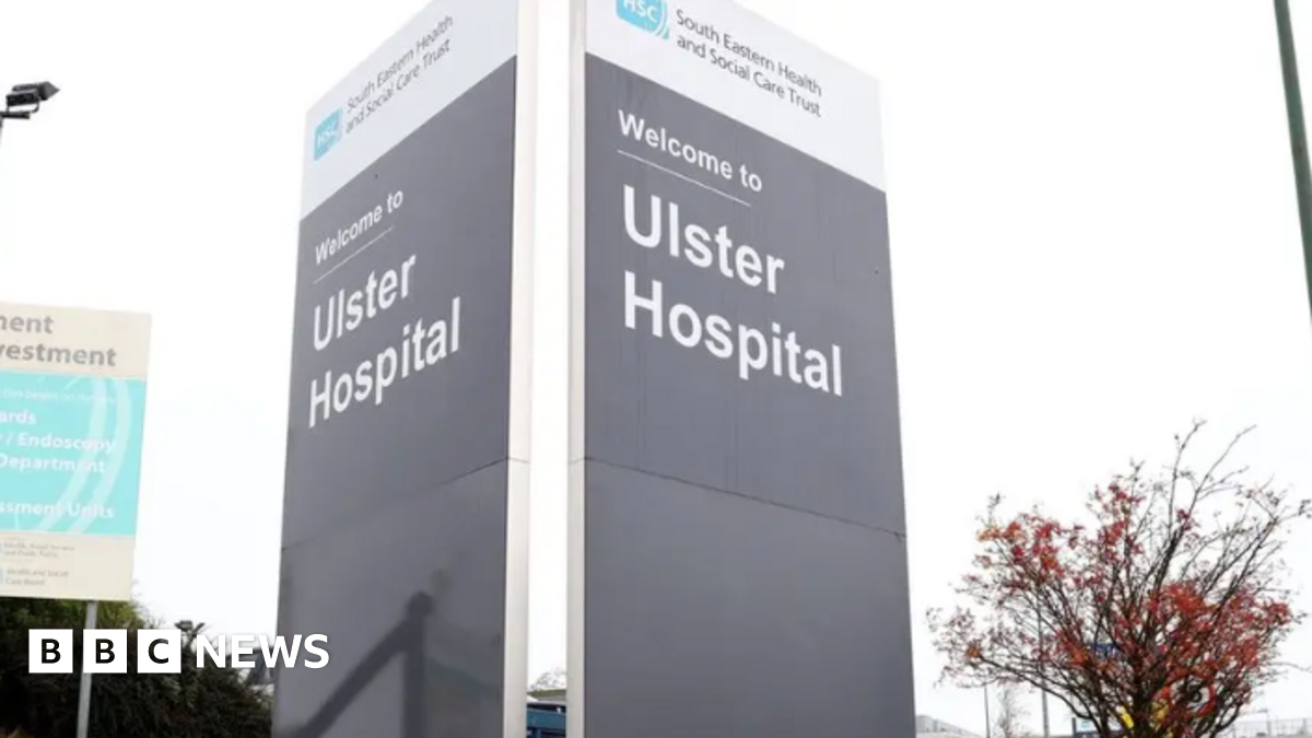 A large grey sign with 'Ulster Hospital' written on it in grey writing. At the top there is a white band with 'South Eastern Health and Social Care Trust' written on it. Next to it is tree with red leaves and another sign post.