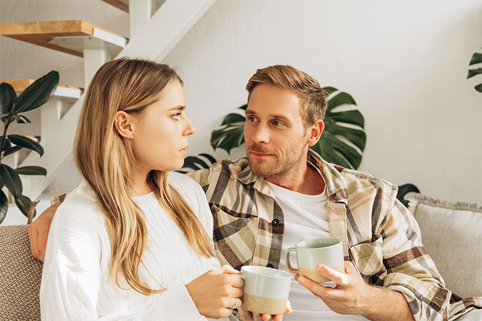 Woman questions marriage while holding coffee cup, looking concerned as husband gazes at her thoughtfully on couch.