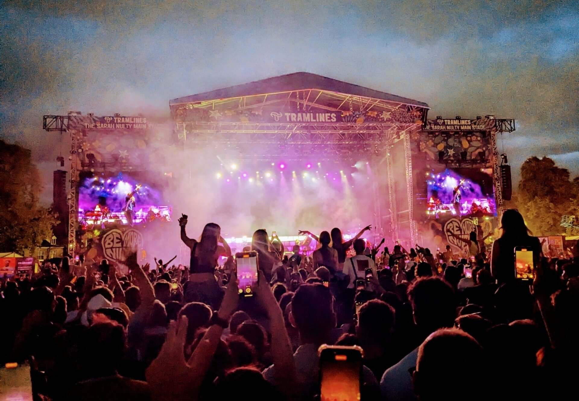 A photo taken at a huge outdoor gig from the back of a crowd watching a band on a massive stage, all lit by pink and purple lighting