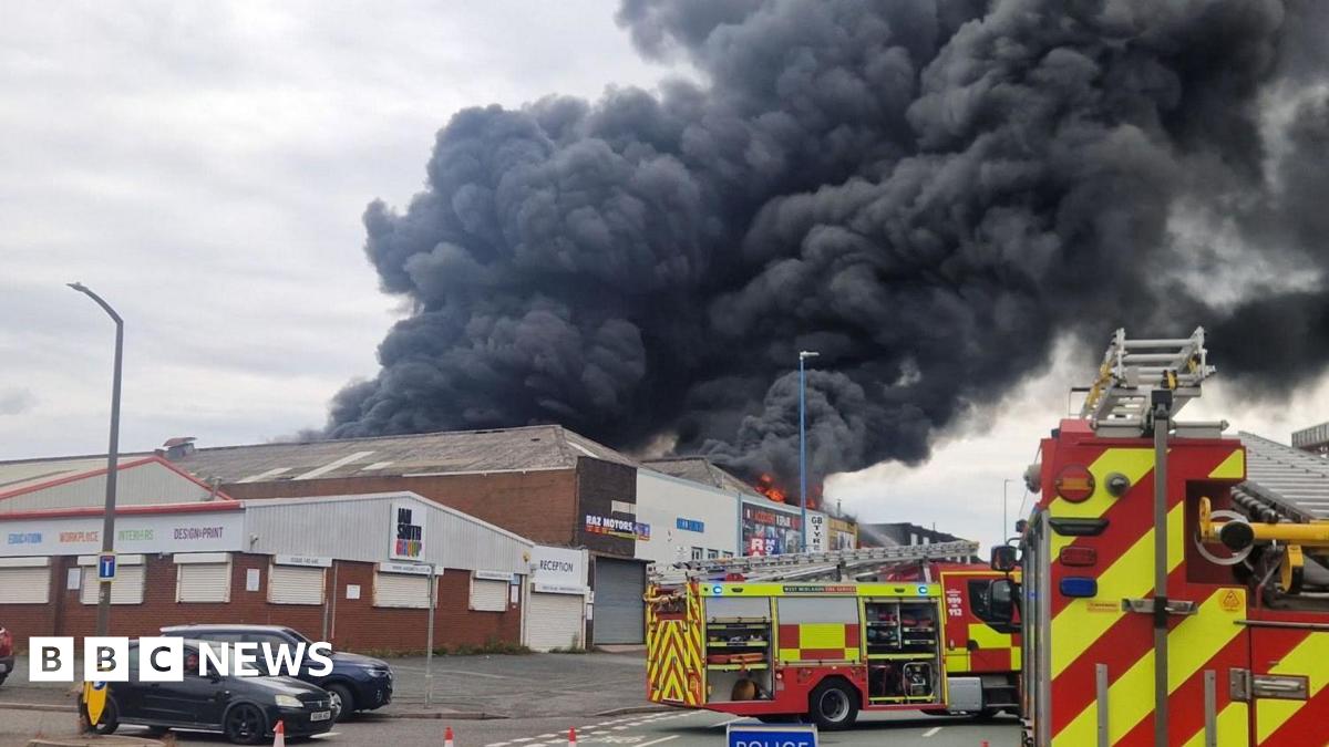 A factory fire has sent great plumes of smoke into the air. Two fire appliances can be seen outside it.