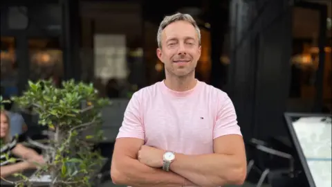 BBC/Ben Moore A man is standing with his arms crossed facing the camera. He has grey hair and is wearing a pink t-shirt and a silver watch. Behind him is a restaurant