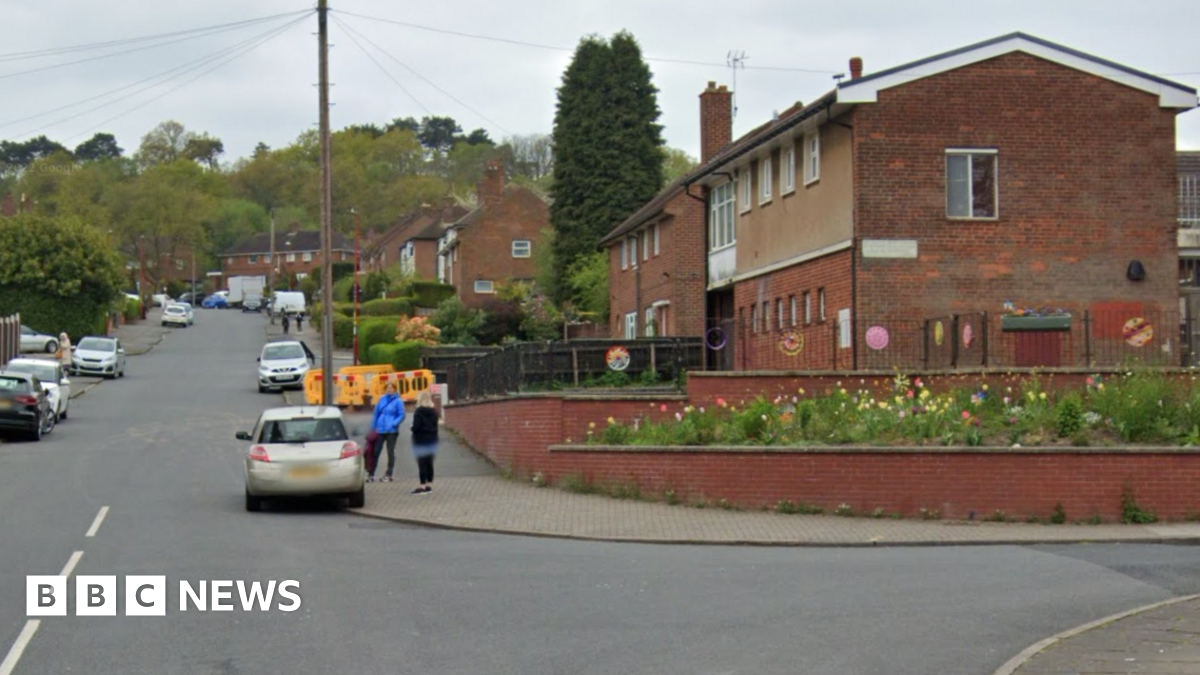 Two storey red brick buildings stretch away from the camera on a residential street. There are cars parked either side of the road, and trees at the top of the street