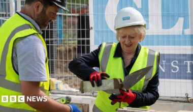 A woman with short blonde hair lays a brick wearing a high-vis yellow vest, hard hat and gloves. A man, also insafety gear, assists her.