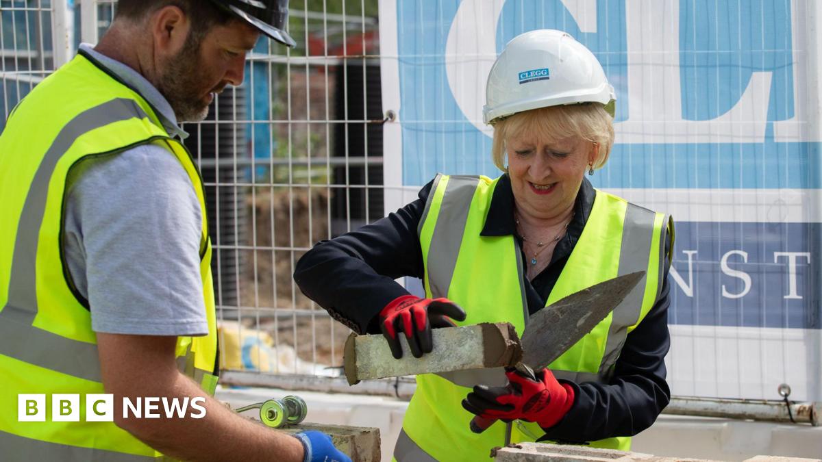 A woman with short blonde hair lays a brick wearing a high-vis yellow vest, hard hat and gloves. A man, also insafety gear, assists her.