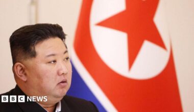Kim Jong Un looking to the side. Behind him is a large North Korea flag.