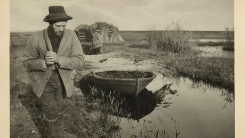 Roseberys Sepia coloured image of a man on a river bank pulling a small boat filled with reeds. 