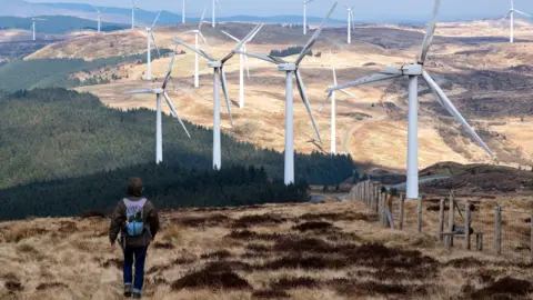 Mike Kemp Wind turbines at Cefn Croes Wind Farm on 16th March 2025 in Cwmystwyth, Wales, United Kingdom. Cefn Croes is a wind farm in Ceredigion, located in the Cambrian Mountains.  A person can be seen walking in the foreground, wearing a dark brown coat and purple backpack.