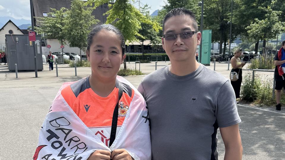 Isabelle stands with her dad Leon. Isabelle is wearing a neon orange top with a flag with writing on it wrapped around her. She has her dark hair tied back. Leon is wearing a dark grey t-shirt and glasses. They are both smiling at the camera.
