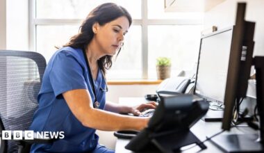 Nurse with long brown hair and blue scrub outfit works at a computer